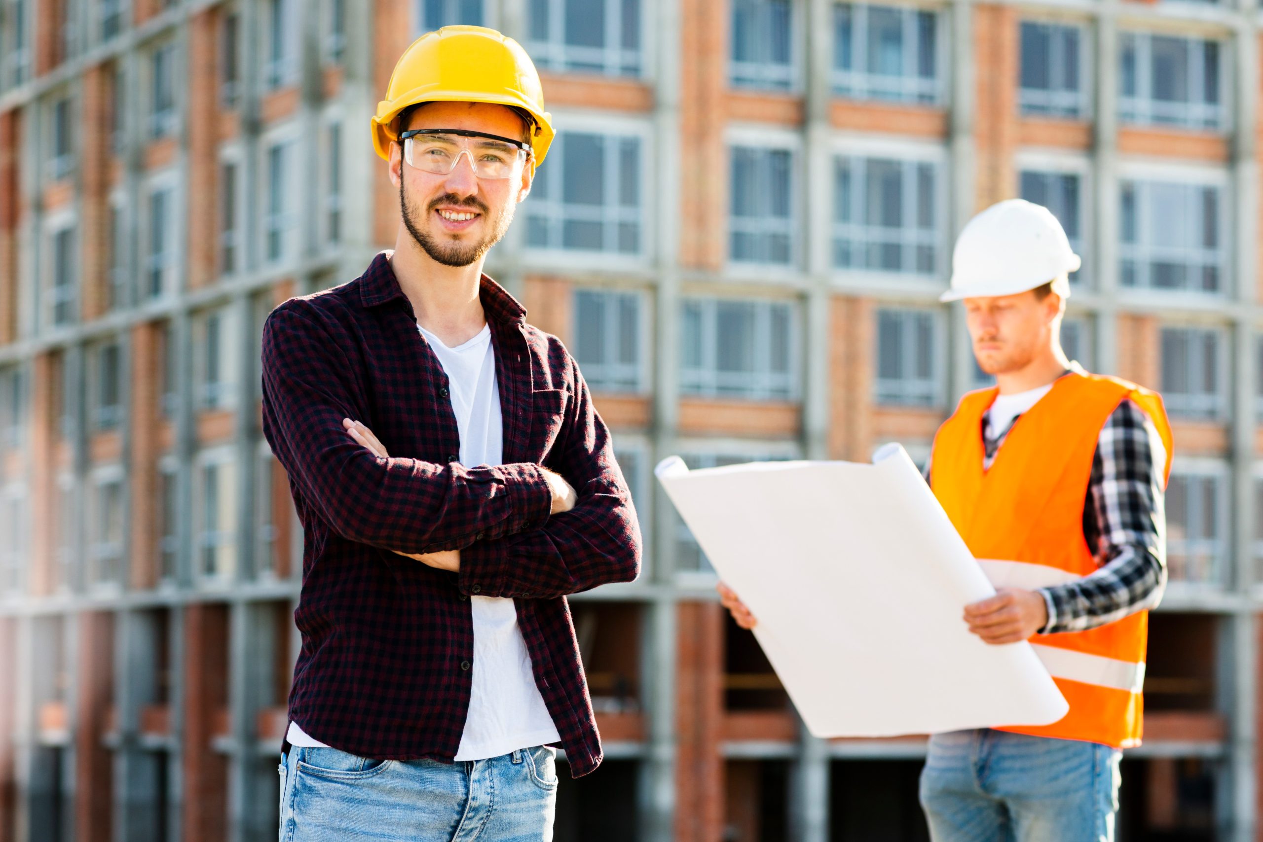 Two construction professionals wearing hard hats stand in front of a building under construction. One person in a yellow helmet and safety glasses stands with arms crossed, while another in a white helmet and orange safety vest reviews large architectural plans.