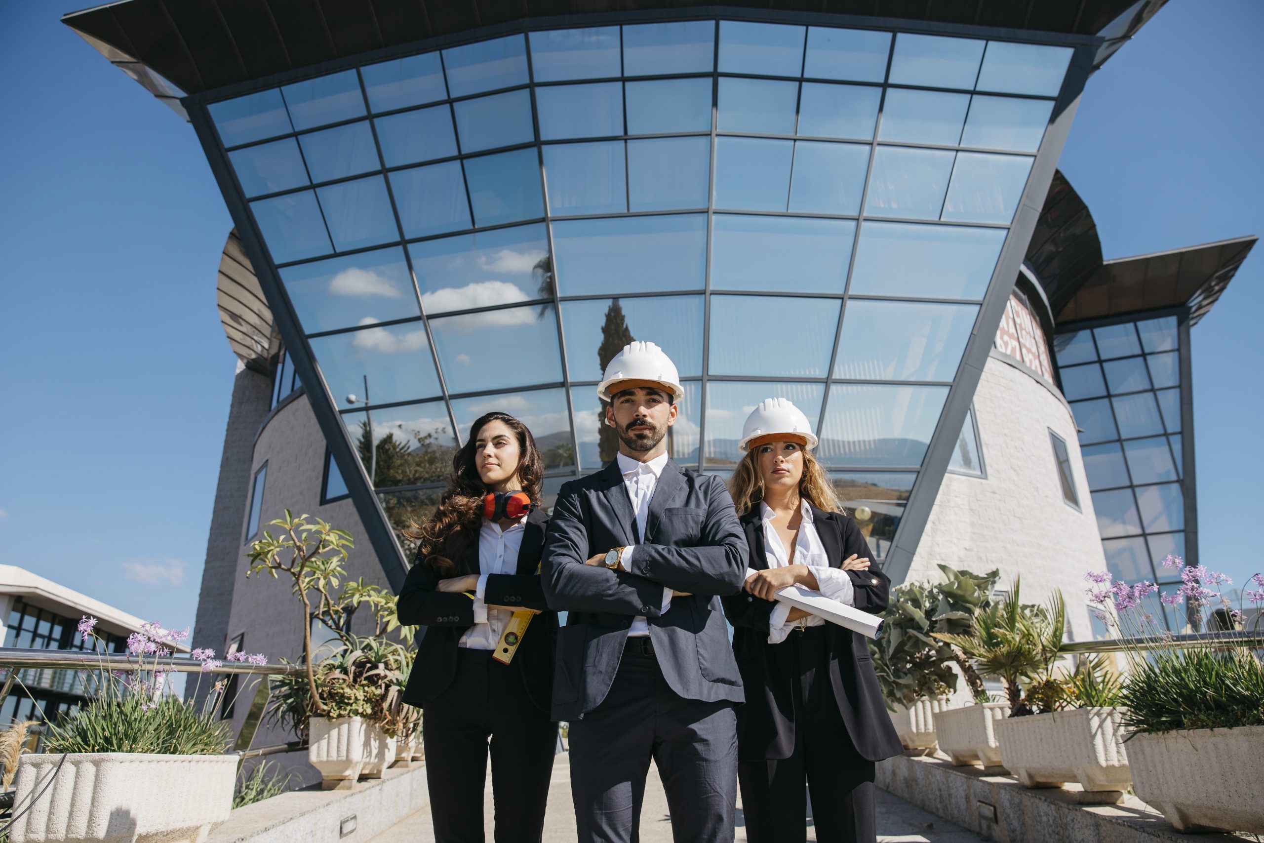 Professional commercial construction consultants standing confidently in front of a modern office building wearing safety helmets and business suits.