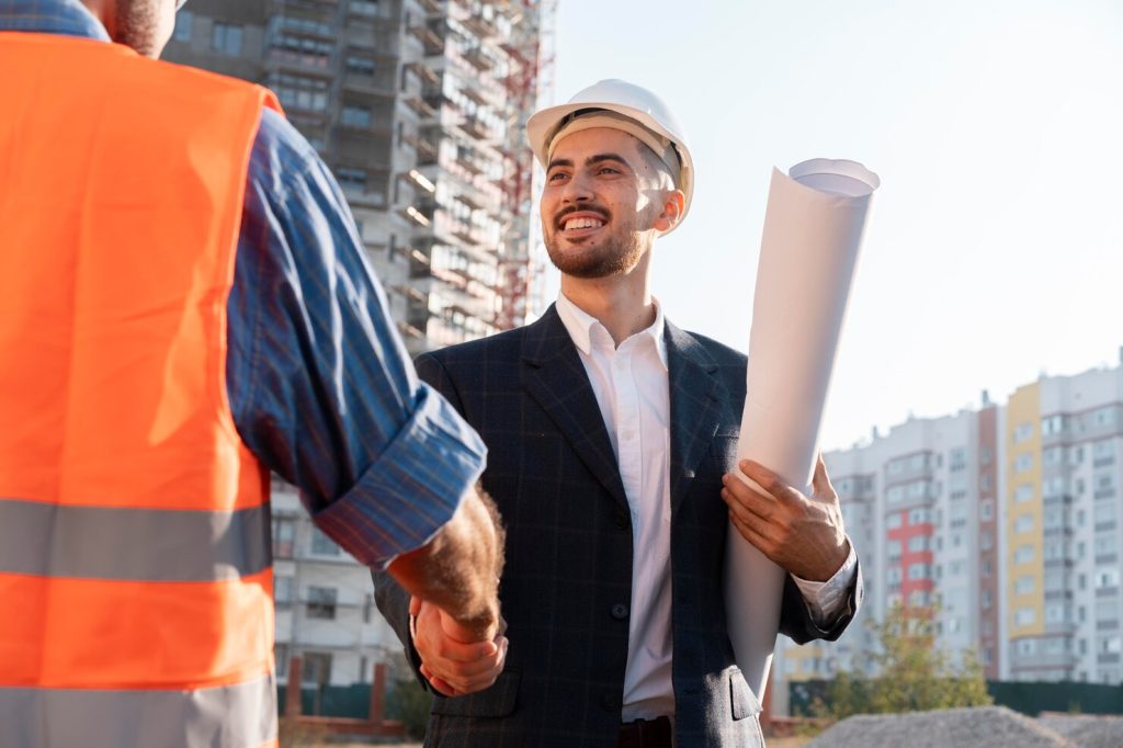 Construction manager in a white hard hat shaking hands with a construction worker wearing a high-visibility safety vest at an urban building construction site while holding rolled blueprints.