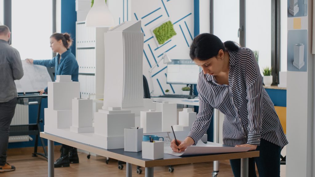 A female architect is working on architectural drawings at a table filled with white-scale building models in a modern design studio, while two colleagues review plans in the background.