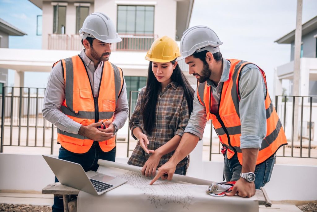 Three construction professionals wearing hard hats and high-visibility vests review architectural plans beside a laptop at a residential building site.