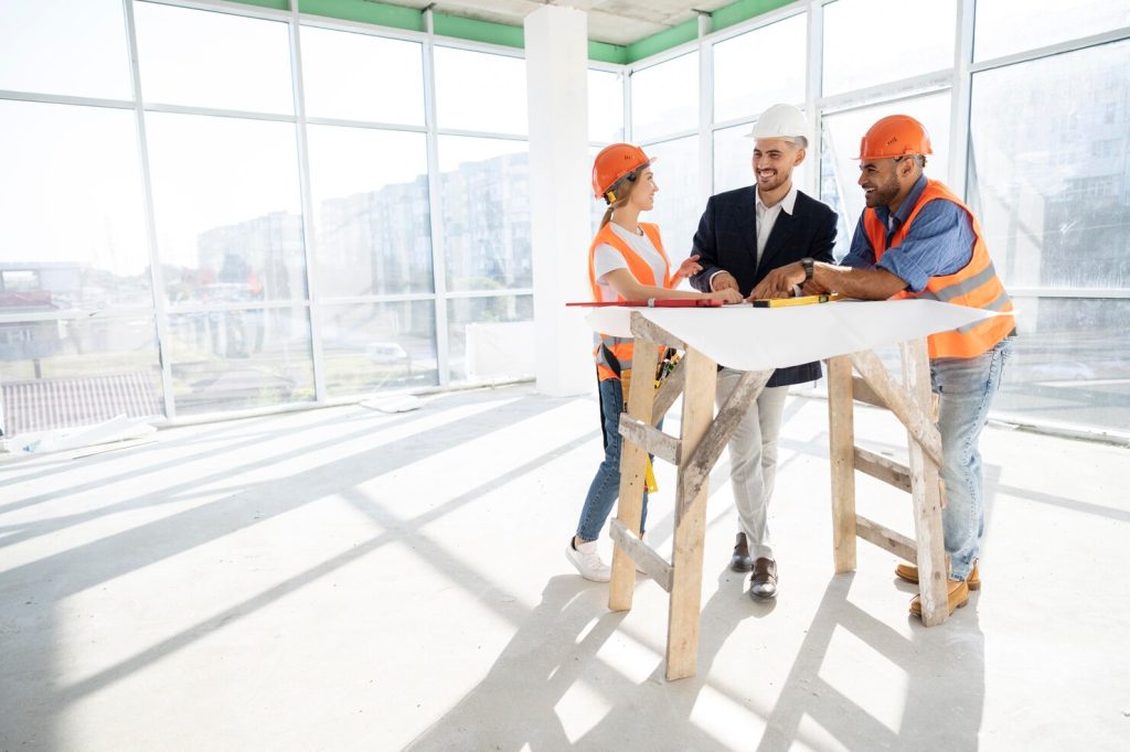 Three construction professionals wearing hard hats and high-visibility vests review building plans inside a bright unfinished office space with large windows.