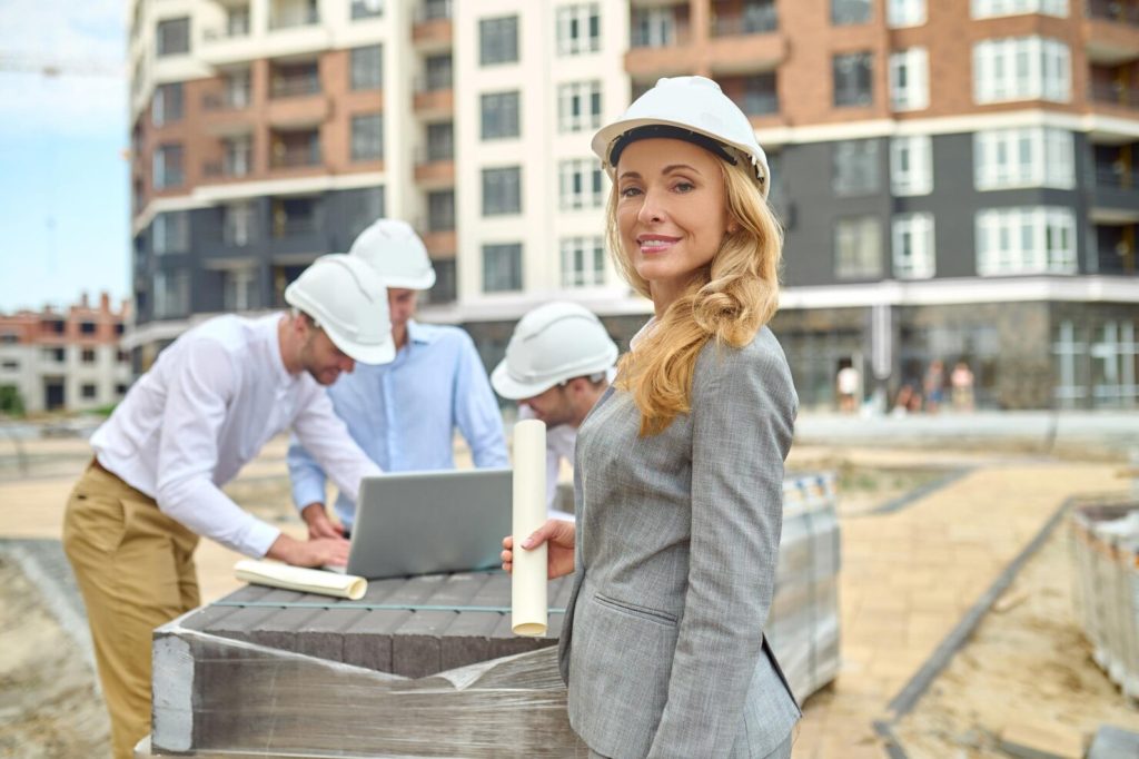 Female construction engineer wearing a white safety helmet holding building plans at a residential construction site with her team in the background.