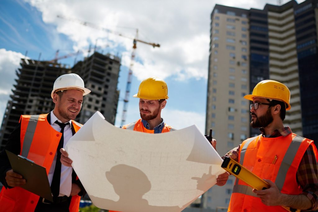 Three construction professionals wearing hard hats and high-visibility safety vests review architectural blueprints at an urban construction site with cranes and high-rise buildings in the background.