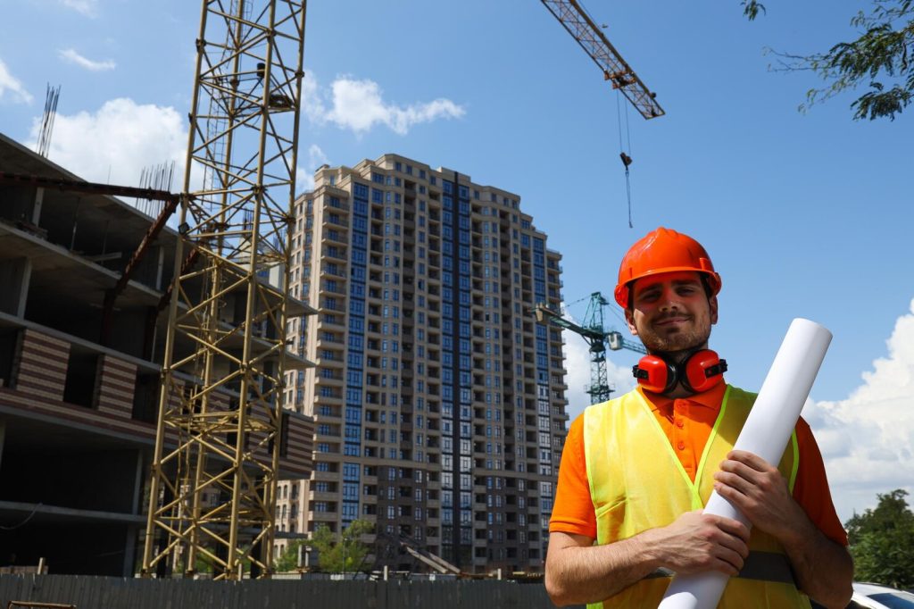 Construction engineer wearing a safety helmet and reflective vest holding blueprints at a high-rise building construction site with cranes in the background.