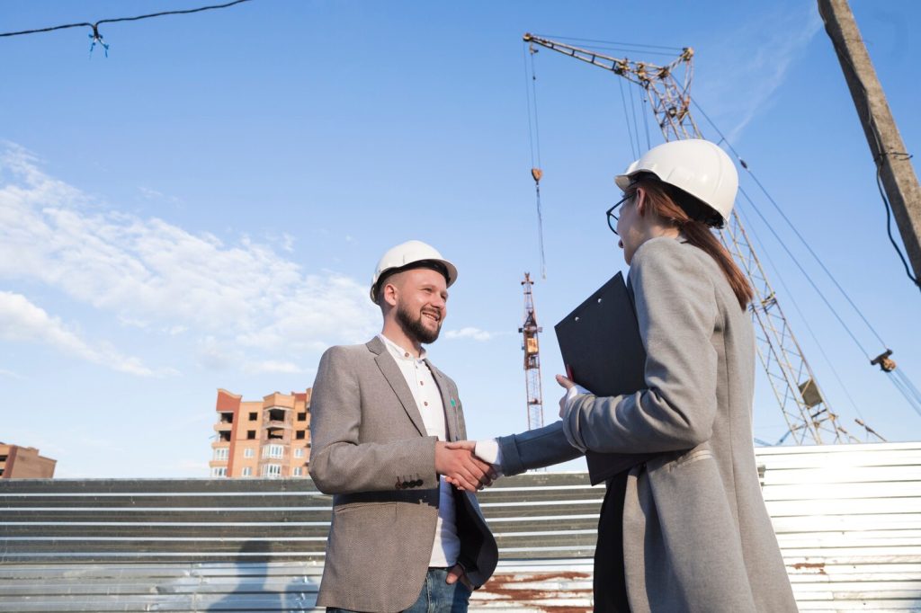 Two construction professionals wearing safety helmets shaking hands at a construction site while discussing revenue planning and project management.
