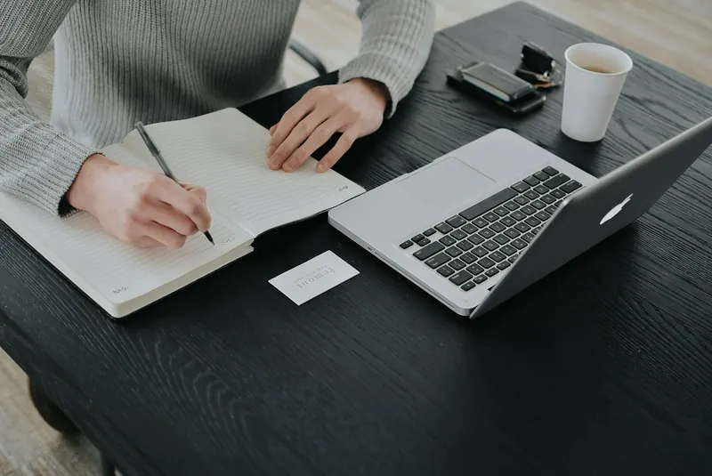 Person writing in a notebook at a desk beside a laptop